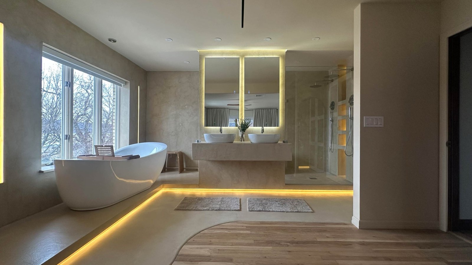 Spa bathroom with a freestanding tub at the window, backlit mirrors and cove lighting